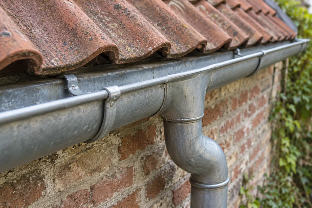 Detail van een zinken dakgoot met bevestigingsbeugels en aansluiting op een regenpijp onder dakpannen